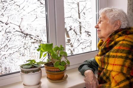 Old lonely woman sitting near the window in his house and looking and dreaming at winter