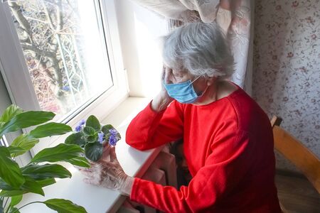 The Covid-19, health, safety and pandemic concept - senior old lonely woman wearing protective medical mask sitting near the window in his house for protection from virusの写真素材