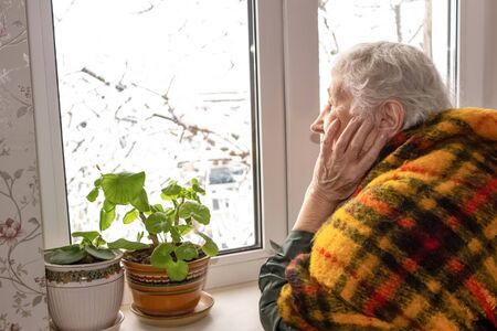 Old lonely woman sitting near the window in his house and looking and dreaming at winterの写真素材