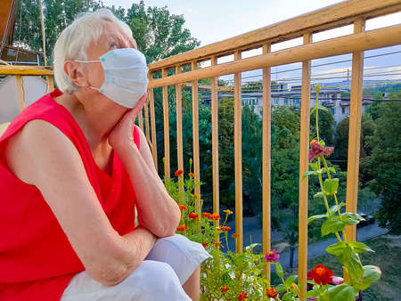 The Covid-19, health, safety and pandemic concept - senior old lonely woman wearing protective medical mask standing at balcony at home for protection from virusの写真素材