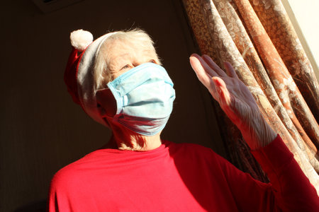 Senior woman near the window in the sunny morning with a Santa hat wears a medical mask due to the . Self isolation away from everyone to avoid contagionの写真素材