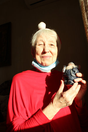 Senior woman near the window in the sunny morning with a Santa hat wears a medical mask due to the . Self isolation away from everyone to avoid contagionの写真素材