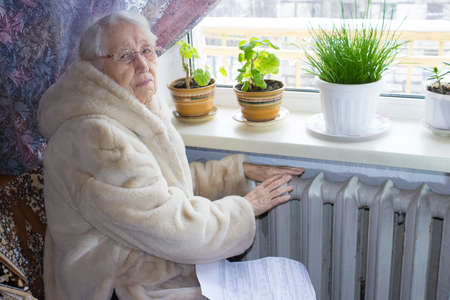 The senior woman holding gas bill in front of heating radiator. Payment for heating in winter.