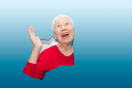 Surprised senior woman looking through hole in paper wall. Shocked lady looks through ripped paper. Sale, discount, season sales. Copy spaceの写真素材