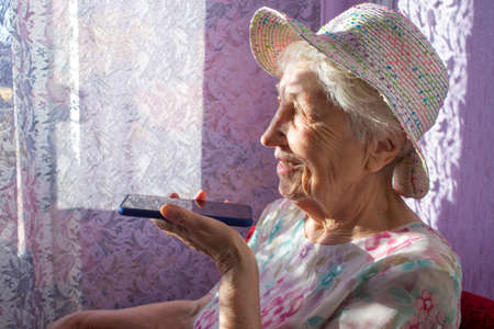 Charming elderly woman in rectangular black rimmed eyewear using voice recognition function on cell phone. Modern senior female holding electronic gadget, sending vocal message via online messengerの写真素材