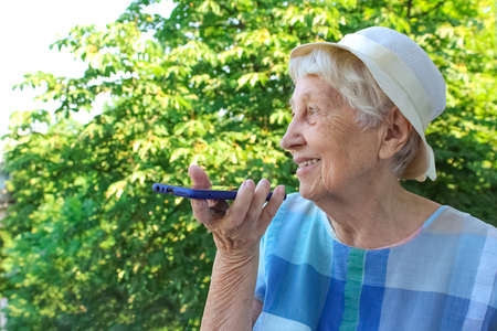 Charming elderly woman in rectangular black rimmed eyewear using voice recognition function on cell...