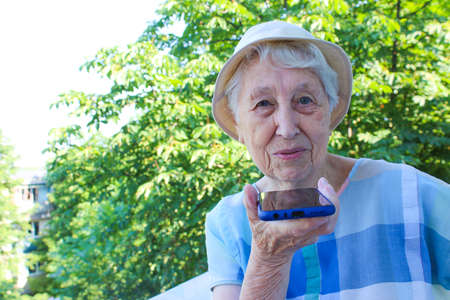 Charming elderly woman in rectangular black rimmed eyewear using voice recognition function on cell...