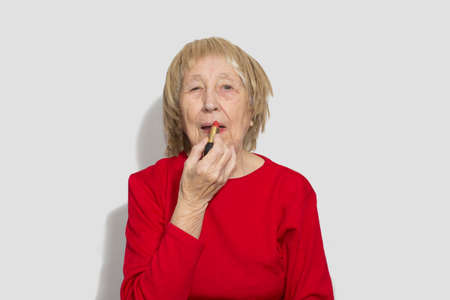 Funny senior woman in wig putting on lipstick on lips isolated on white studio background