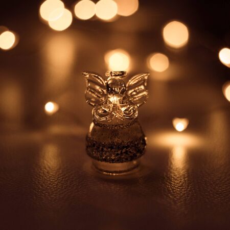 A small transparent angel with wings holds a star on a dark brown background with bright lights bokeh. Glass angel close-up, macro. Christmas still life. Beautiful shiny bokeh.の写真素材