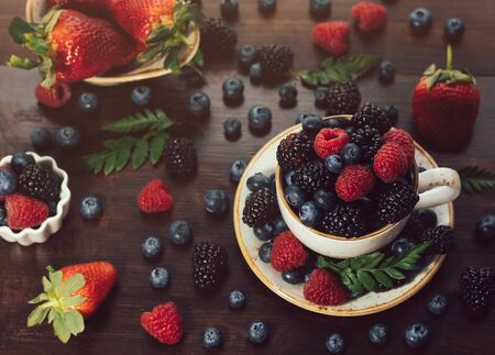Tea cup with berries on a dark wooden background. Raspberry, blackberry, bilberry in a cup. Beautiful juicy bright berries. Summer berry mix.の写真素材