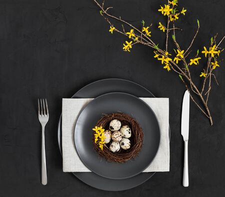 Easter table setting with a nest of quail eggs , a napkin and yellow flowers on a black background....