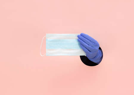 A female hand in a blue glove holds a medical mask on a pink background. Hand in a paper hole. Prevention of coronavirus, covid-19. Healthcare concept.の写真素材