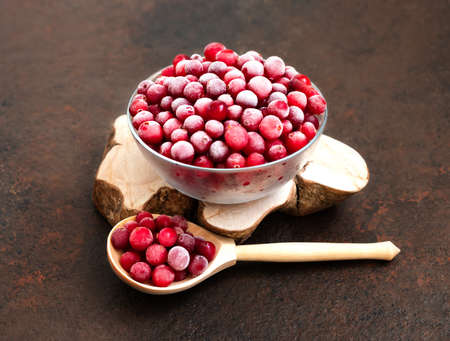 Frozen cranberries in a bowl and wooden spoon on a brown background. Frozen healthy food. Side view.