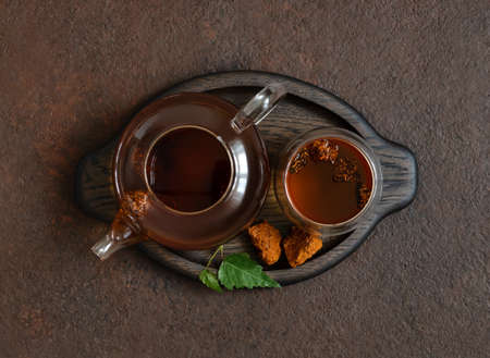 Chaga tea in a glass bowl and teapot on a brown background. Organic infusion with chaga mushrooms in a wooden tray. View from above. Healthy beverage.の写真素材