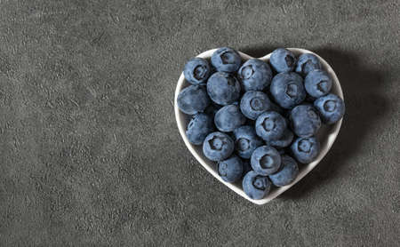 Blueberries in a heart shaped plate on a gray background. Fresh juicy blueberries. Healthy food concept. Top view, flat lay, copy space.の写真素材