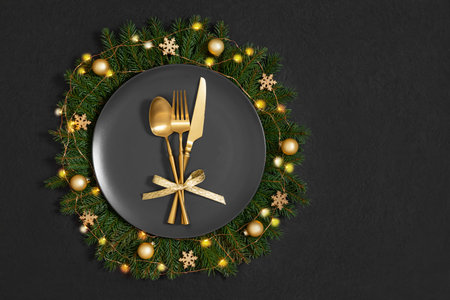Black plate in a Christmas wreath with gold cutlery and decor on a black background. Festive table setting. Copy space. Top view, flat lay.の写真素材