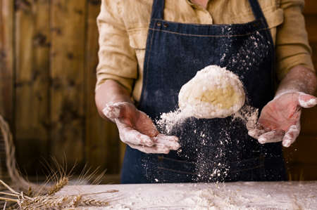 Womens hands, flour and dough. Levitation in the frame. A woman in an apron is preparing dough for home baking. Rustic style photo. Wooden table, wheat ears and flour in the crucifix. Emotional photoの写真素材
