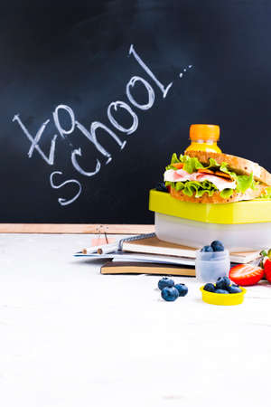 Box with school lunch near the black board. Healthy food for a child. Toast with salad and strawberries and blueberries and a bottle of juice. Food and books for the institute or school. Copy space.の写真素材