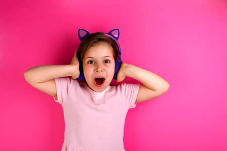 Little girl in headphones listens to music. European child on a pink background is having fun. Free...