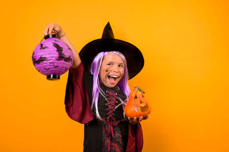 Happy Halloween! Cute cheerful little witch in a carnival costume and a pumpkin in her hands. A beautiful childish girl on a yellow background conjures and laughs. Selective focus. High quality photoの写真素材