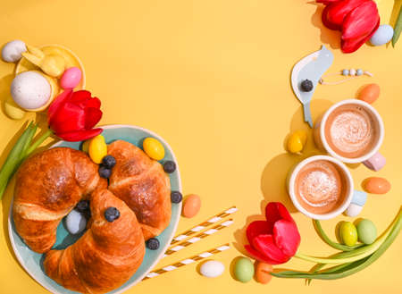 Spring breakfast with croissants and berries. Two Espresso coffee in bright cups, Easter chocolate eggs, pink bunny ears and red tulips on a yellow background. Copy space. Flat layの写真素材