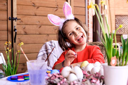 Cheerful crazy girl in rabbit bunny ears on headat home at the table with easter decoration, Paints eggs. Easter painting eggs on eyes. Easter and holidays concept. Copy space.の写真素材