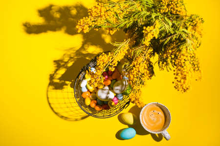 A lot of multi-colored Easter eggs, a mimosa branch and a mug of aromatic coffee on a yellow background. Happy Easter card with copy space for text. Minimal style. In a hard light. selective focusの写真素材