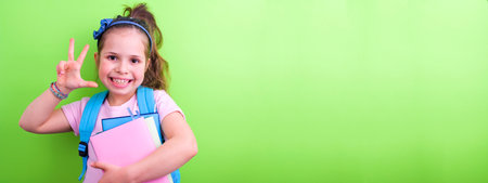 Schoolgirl with a backpack and books with colored covers on a green background. The little girl is happy and happy, ready to go to school. Great place for text. Bann, long formatの写真素材