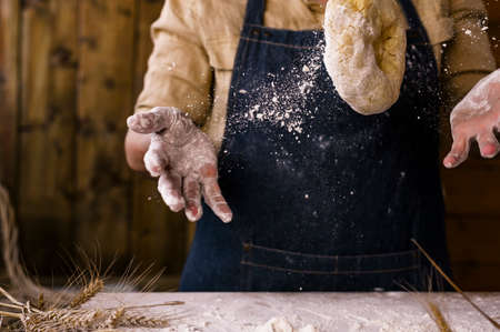 Womens hands, flour and dough. Levitation in a frame of dough and flour. A woman in an apron is preparing dough for home baking. Rustic style photo. Wooden table, wheat ears and flou.Emotional photoの写真素材