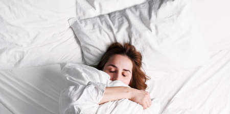 Girl wakes up in the morning. A young woman in bed hugs a pillow snugly and comfortably. Shows no desire to wake up. White bed, blanket and pillows. Free space for text. Copy space. Bannerの写真素材