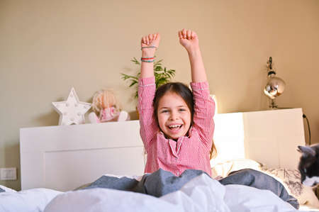 The little girl woke up. The child in his pajamas stretches in bed, smiling shows positive emotions. Morning to school. Copy spaceの写真素材