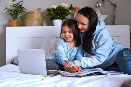 Distance learning online education. Schoolgirl with digital tablet laptop notebook and doing school homework. Mom does homework with her daughter at home in bed. Interactions of children and parents.の写真素材