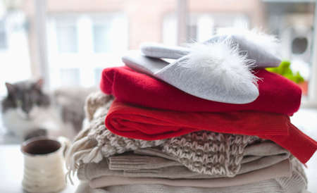 stack of cozy knitted sweaters and slippers hom. near the big window. On the background sits a...