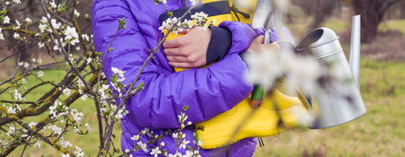 The girl holds bright yellow rubber boots, a watering can, a spade in her hands. The flowering...