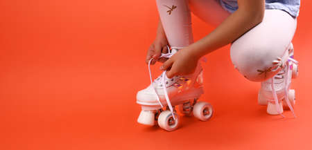 Little child with roller skates, a blue helmet on a red background, tying shoelaces. A girl of 7 years old poses and prepares for active leisure on retro ice skates. Copy spaceの写真素材