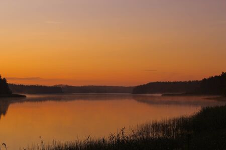 orange dawn on the lake in the forest with fog. Amazing orange sunrise on a lake with sunshine, trees and reflections in the waterの写真素材