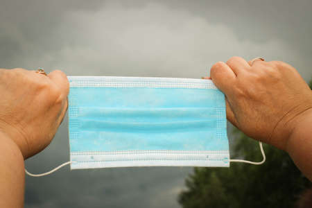 Woman's Hands holding blue medical protective mask on a dark cloudy Background. Concept of social...