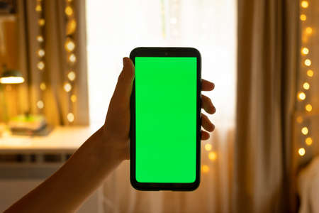 Smartphone with Green Mock-up Screen. Green Chroma Key Screen Device Phone is in a Teen Hand in the Kid Room. In the Background Cozy Home with Christmas Decoration in the Evening with Warm Lights onの写真素材