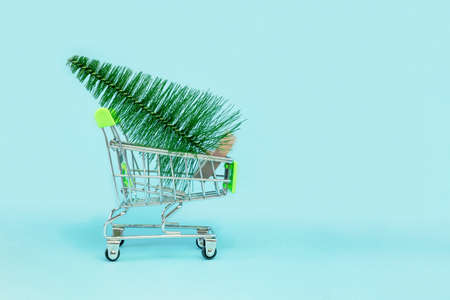 Small Christmas tree is in a shopping cart. Artificial mini Christmas trees in a supermarket trolley on blue background. Christmas, New Year, Holiday shopping concept. Minimalismの写真素材