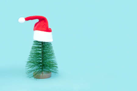 Small Christmas tree in red Santa Hats. Decorating of Artificial Fir tree. Beautiful imitation of a mini Christmas tree, with Santa Cap on blue background. Christmas holidays concept. Minimalismの写真素材
