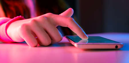 Use of mobile phone in trendy neon lights. Creative vivid color of ultraviolet red and blue. Hands of Teen Girl scrolling up photos Close-up at dark neon room. App for sale in shop, Social media.の写真素材