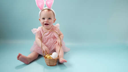 Happy baby girl, ten months old, wearing bunny ears headband, sitting on light blue studio background and playing with colored easter eggs. Little Child. Easter greeting card. Baby easter bunny, 4Kの写真素材