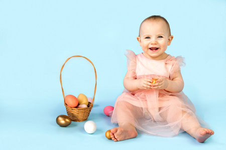 Happy baby girl, ten months old, wearing bunny ears headband, sitting on light blue studio background and playing with colored easter eggs. Little Child. Easter greeting card. Baby easter bunnyの写真素材