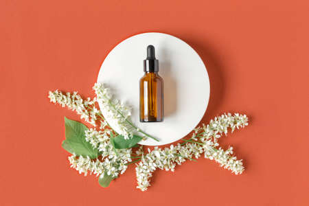 Amber glass dropper cosmetic bottle on oval podium, red orange background. Natural skin care, SPA design. Mineral organic oil. Product Mock-Up. Oily pipette. Flat lay, Top View. White flowersの写真素材