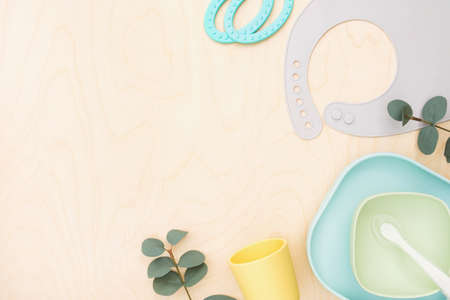 Baby accessories and tableware for food on wooden background. First feeding for kids concept. Flat lay, Top View, Space for text. Silicone dishware, bib, cup on neutral backdrop. Serving baby foodの写真素材