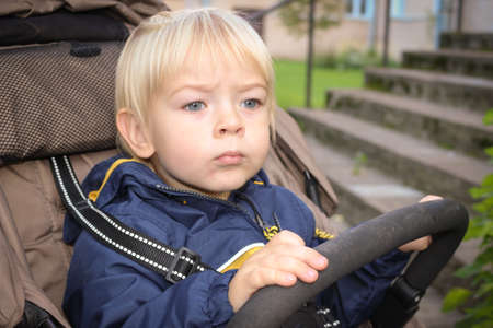 Very beautiful blonde hair boy with blue eyes sitting in the pram outdoor. Cute little child in a stroller walking through autumn park. child in buggy. Kid in a pushchair. Traveling with children.の写真素材