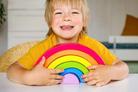 Little blond hair boy in clothes made of natural fabric plays with rainbow colored wooden toys at...