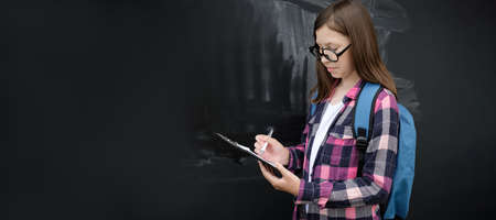 Teenage student girl in shirt, glasses with blue backpack and diary or notebook over black chalkboard background. Education, school and people concept. Teen girl with book near blackboard. copyspace.の写真素材