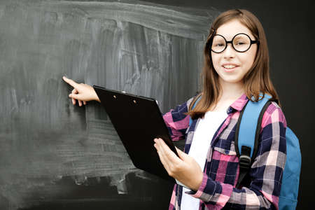 Teen girl finger pointed to blackboard. Teenage student girl in shirt, glasses with blue backpack...