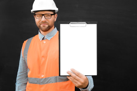 Happy Engineer Pointing At Blank Paper Sheet, Clipboard. Man Builder In Safety Vest And Hard Hat Advertising Empty White Tablet Standing Over Black Background, Mockup. All For Construction. copy spaceの写真素材